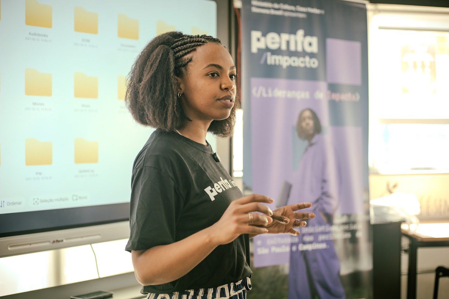 Jovem negra de tranças, com camiseta do Perifa Impacto, apresentando diante de um telão numa formação da Casa Hacker