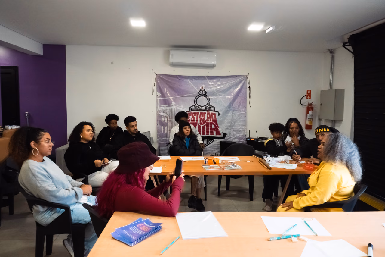 Participantes de uma atividade dos Hubs de Cidadania Ativa conversam e acompanham uma dinâmica coletiva em torno de uma mesa. Ao fundo, uma faixa com a identidade visual da atividade ocupa parte da parede.