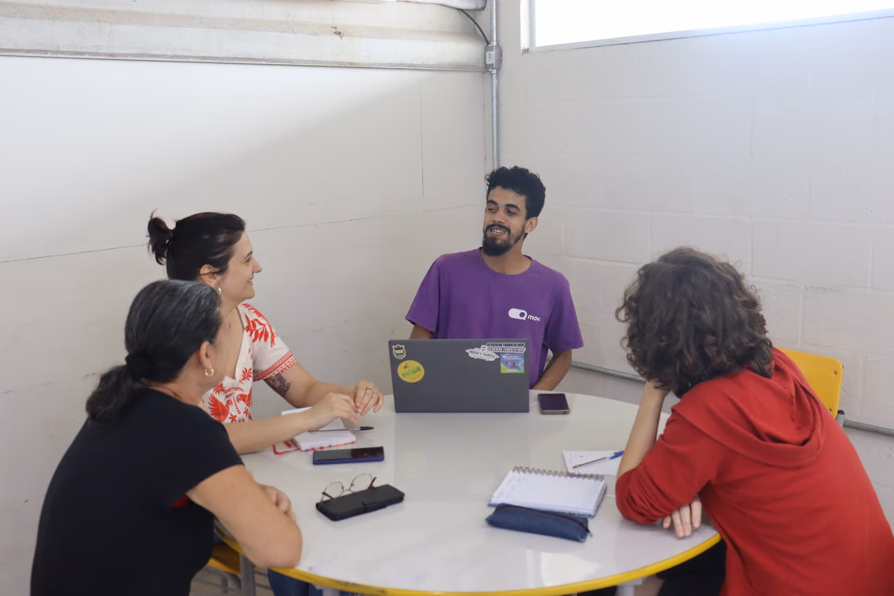Quatro pessoas conversam em volta de uma mesa redonda com notebook, cadernos e celulares, numa sala de paredes brancas.