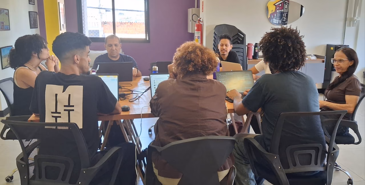 Grupo de sete pessoas reunidas ao redor de uma mesa em uma sala de trabalho. Os participantes utilizam notebooks e acompanham uma atividade coletiva, enquanto conversam e compartilham informações.
