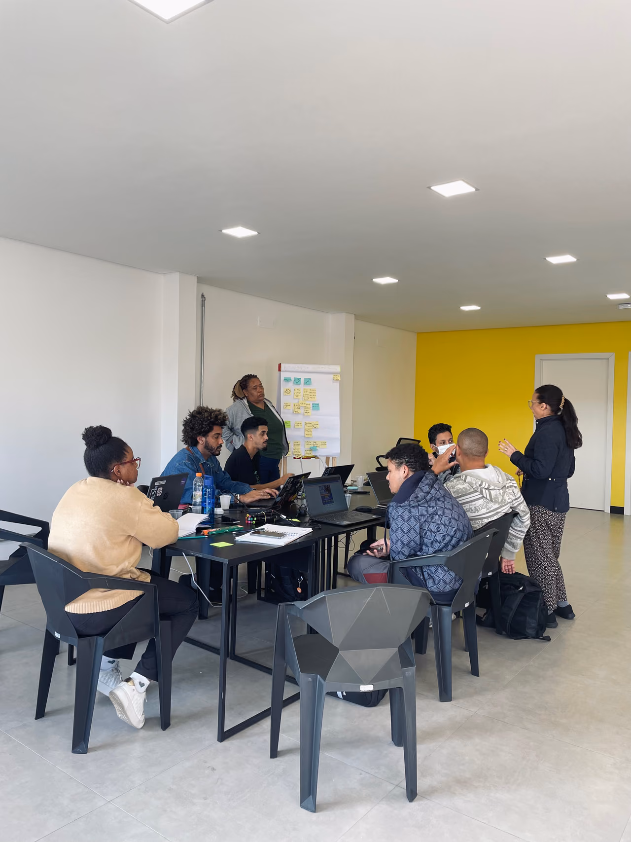 Participantes de uma formação dos Hubs de Cidadania Ativa trabalham em grupo com notebooks, enquanto uma facilitadora conduz a atividade. Ao fundo, há um quadro com anotações e materiais utilizados durante a dinâmica.