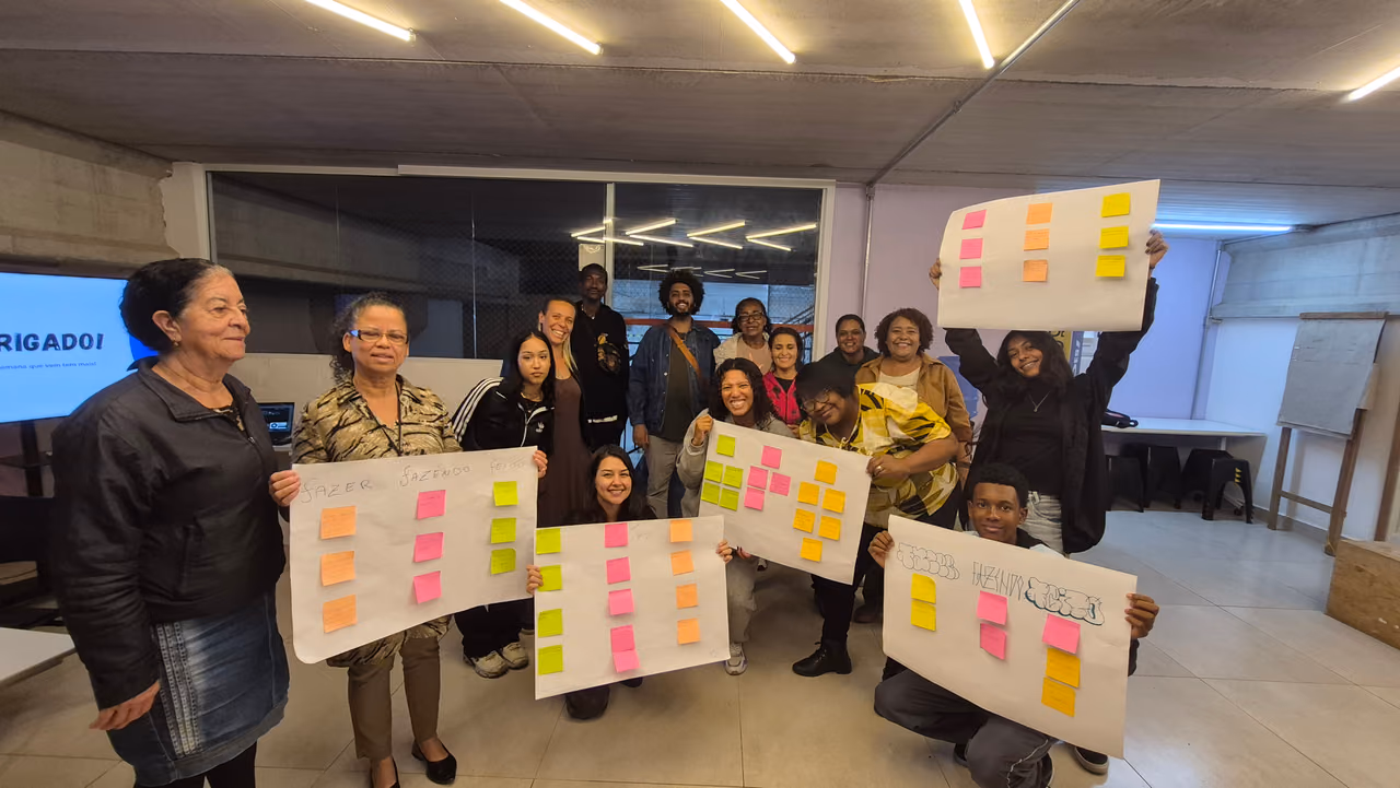 Participantes de diferentes idades reunidos durante uma atividade formativa dos Hubs de Cidadania Ativa. Algumas pessoas seguram cartazes com post-its coloridos organizados a partir de uma dinâmica de construção coletiva.