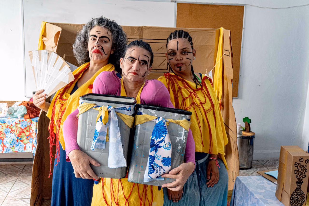 Três pessoas posam diante de um cenário de papelão. Elas usam figurinos coloridos em amarelo, rosa e azul, com pinturas faciais que simulam expressões de palhaços. Duas delas seguram recipientes metálicos decorados com tecidos, enquanto a pessoa à esquerda segura um leque branco.