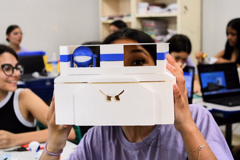 Estudante segura diante do rosto um protótipo branco com detalhes azuis, construído durante uma atividade do Hackerclubes. Ao fundo, outros estudantes trabalham em computadores e materiais de experimentação.