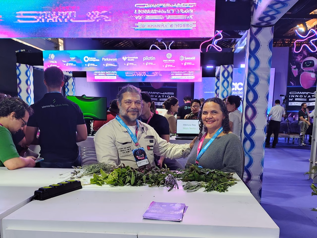 Fátima e Daniel, da Horta do Itajaí, posam lado a lado em um espaço do Campinas Innovation Week 2026. À frente deles, há uma mesa com diferentes tipos de folhas e ervas. Ao fundo, aparecem estandes, telas e elementos coloridos do evento.