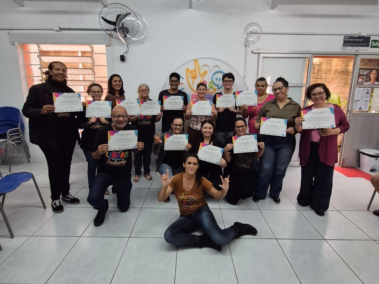 Grupo de alunos e integrantes do projeto posa para uma foto em uma sala. As pessoas seguram certificados de conclusão das aulas de Libras, enquanto uma participante está sentada no chão à frente do grupo fazendo sinais em Libras.