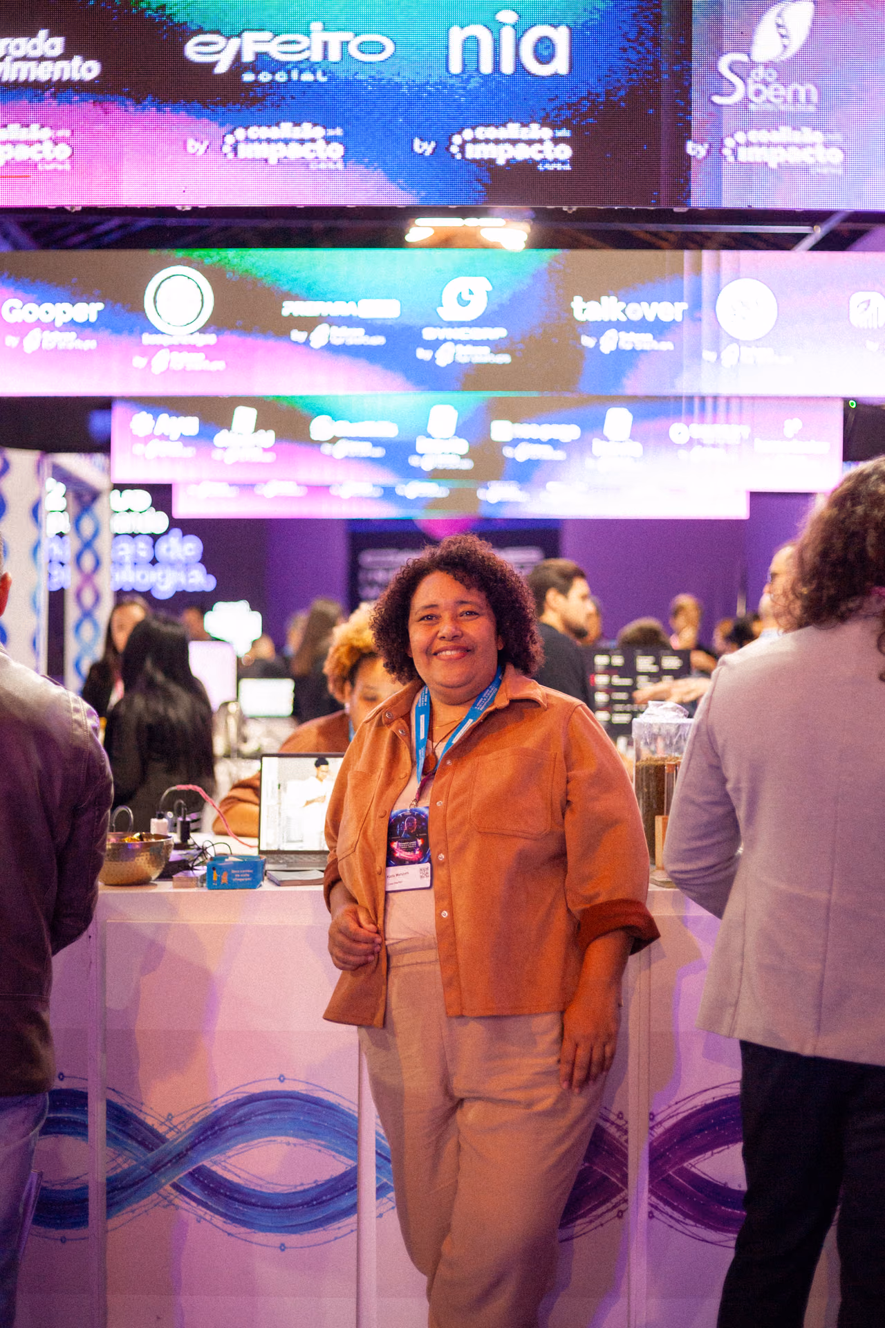 Karla Marques posa para uma fotografia em frente a um balcão de exposição durante o Campinas Innovation Week 2026. Ela veste uma camisa em tom terracota, calça bege e crachá do evento. Ao fundo, aparecem painéis coloridos com as marcas de organizações e iniciativas participantes.