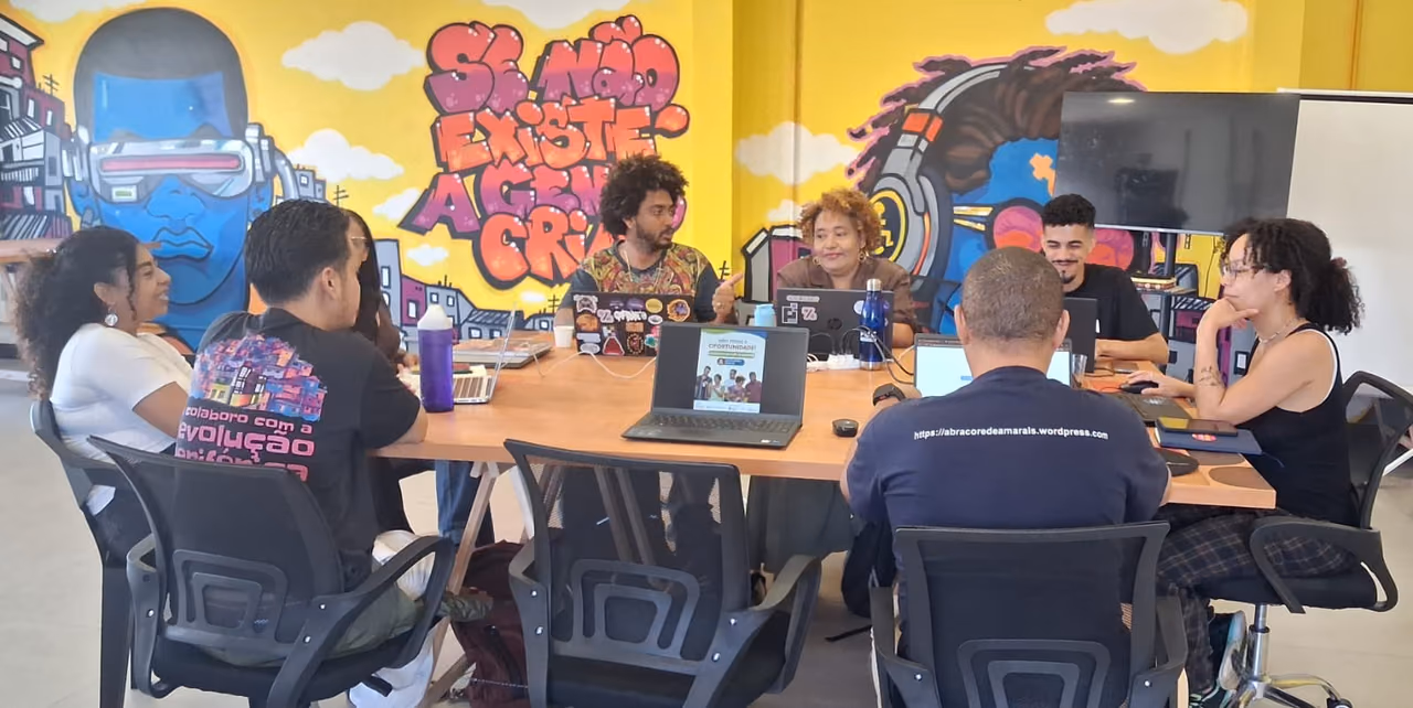 Grupo de pessoas participa de uma reunião ao redor de uma mesa com notebooks e materiais de trabalho. Os participantes conversam em um ambiente com paredes decoradas por grafites coloridos, incluindo referências à cultura e à identidade negra.
