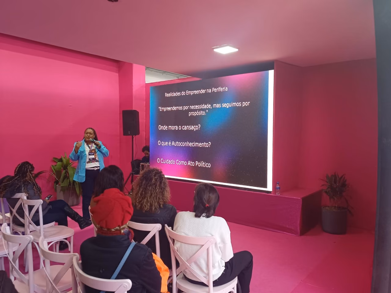 Karla Marques fala ao microfone durante uma oficina, diante de um público sentado. Ao fundo, uma tela apresenta o tema “Realidades do Empreender na Periferia” e tópicos sobre propósito, autocuidado e autoconhecimento.