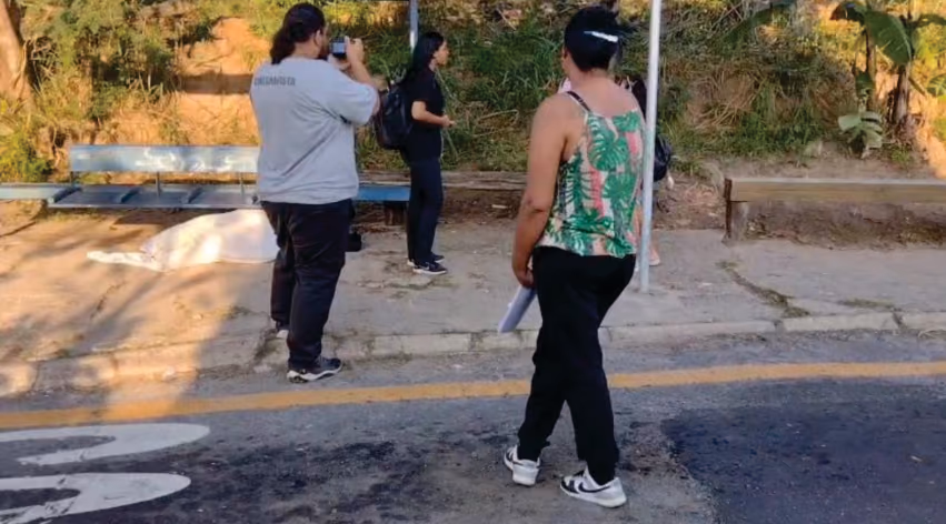 Street scene recording next to a bus stop: one person films with the camera, another is on the scene next to the bench, and a third watches while holding papers.