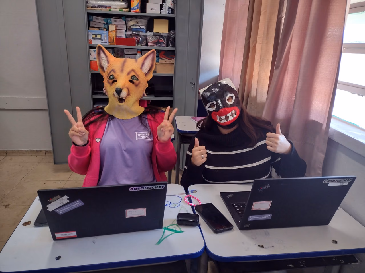 Duas participantes do Minas em Tech estão sentadas diante de computadores em uma sala. Elas usam máscaras de raposa e de personagem, fazem gestos para a câmera e têm dois notebooks sobre as mesas, identificados com adesivos da Casa Hacker e do Minas em Tech.