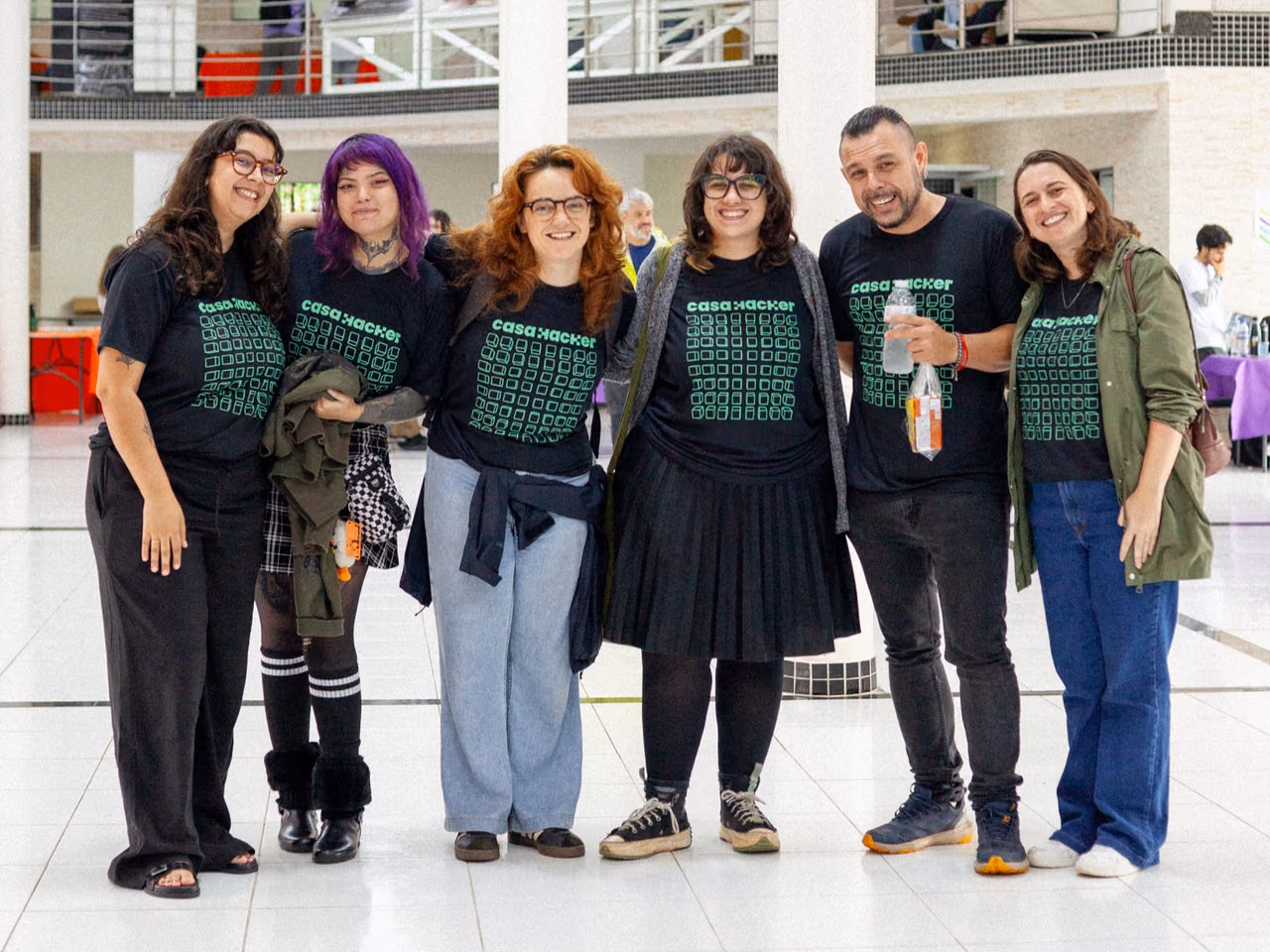 Seis integrantes da Casa Hacker posam juntos no espaço da II Feira de Ciências InterCOCEN. Eles usam camisetas pretas com a identidade visual da organização e estão em um salão com estandes e participantes do evento ao fundo.