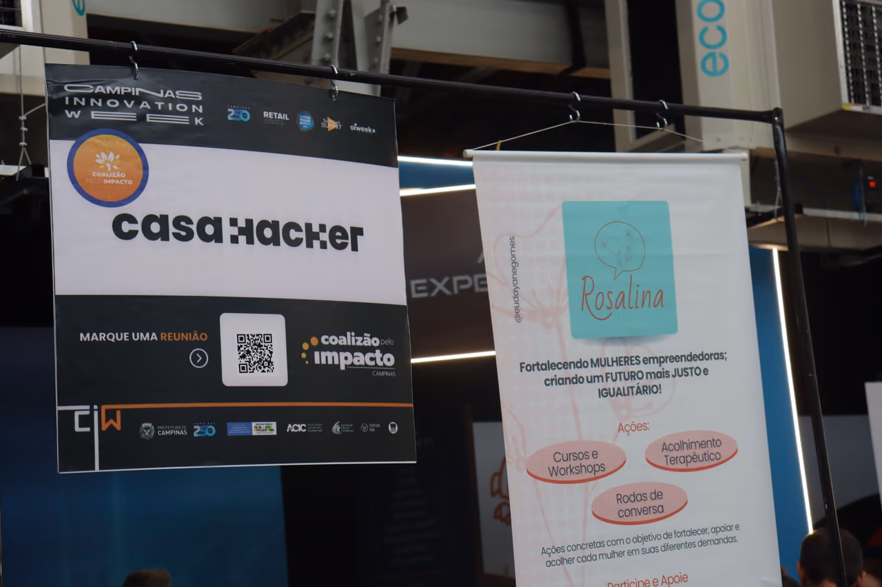 Dois banners suspensos em um espaço do Campinas Innovation Week 2024. À esquerda, um banner da Casa Hacker com a identificação da Coalizão pelo Impacto Campinas. À direita, um banner do Movimento Rosalina apresenta ações voltadas ao fortalecimento de mulheres empreendedoras, como cursos, workshops, acolhimento terapêutico e rodas de conversa.
