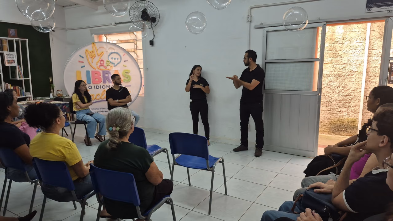 Janaína, colaboradores e alunos participam de uma aula de Libras em uma sala. Duas pessoas estão à frente do grupo realizando sinais em Libras, enquanto os demais participantes acompanham sentados em círculo. Ao fundo, há um painel com a identificação do projeto Libras.