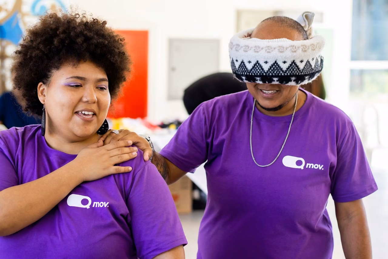 Dois participantes de uma atividade interativa vestem camisetas roxas com o logotipo ‘Q Mov.’. A pessoa à esquerda, uma mulher com cabelo crespo volumoso e maquiagem roxa, sorri enquanto guia a pessoa à direita, que usa um gorro puxado sobre os olhos como uma venda. Ela apoia a mão no ombro do colega, que sorri enquanto segura suavemente seu ombro. O ambiente parece ser um espaço interno iluminado, possivelmente um evento ou oficina.