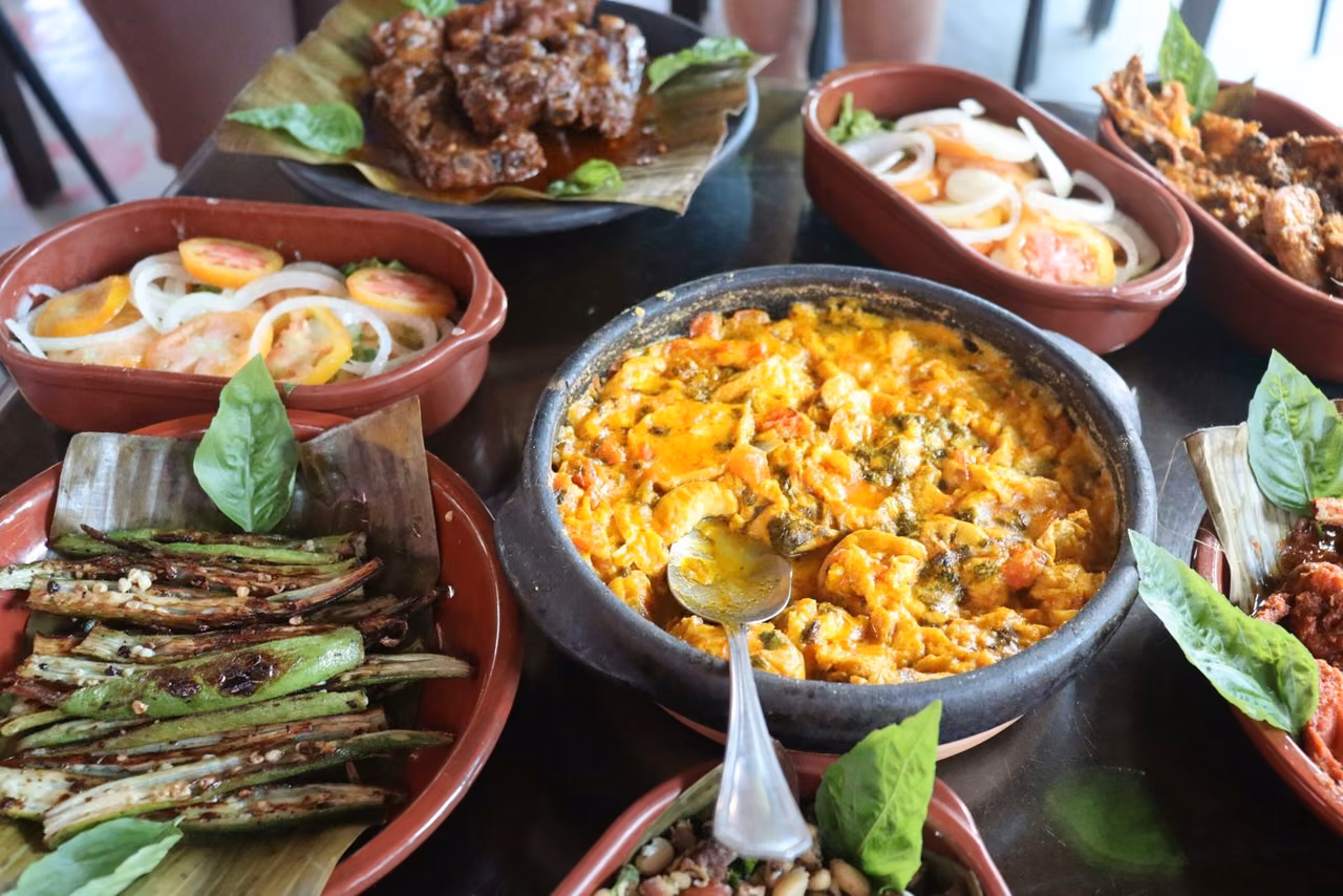 Mesa posta com destaque para panela de barro escuro com moqueca de caju dentro. A moqueca tem uma cor amarelada e a panela está com uma colher dentro.
