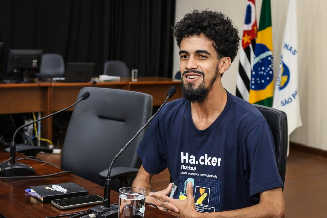 Homem pardo, de cabelo preto cacheado e cavanhaque, vestindo camisa azul com escrita Casa Hacker.